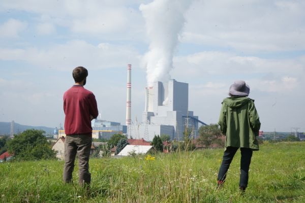 Zwei Personen sind von hinten zu sehen. Sie schauen aus der Entfernung auf ein Kraftwerk. Beim Kraftwerk kommt Rauch aus dem Schornstein