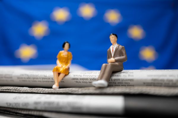 Foto: Zwei Miniaturfiguren sitzen auf einer Zeitung, die Flagge der Europäischen Union ist im Hintergrund zu sehen