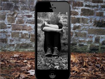 Auf dem Bildschirm eines Smartphone ist in schwarz-weiß ein offenbar trauriger Mensch zu sehen, der am Boden kauert. Rechts und links des Smartphones geht der Hintergrund in  Farbe weiter.